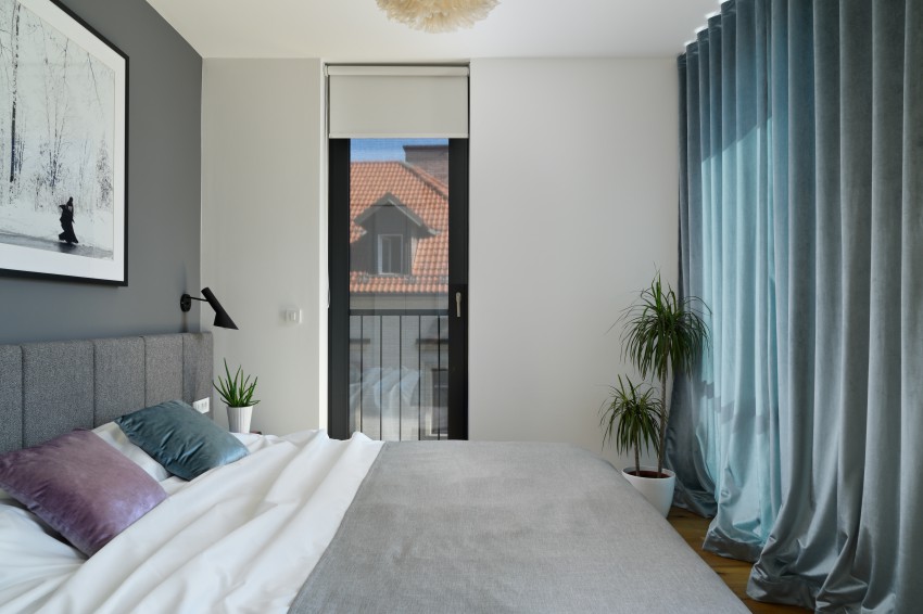 Spalnica je zasnovana minimalistično. Sivo steno in posteljo dopolnjujejo bogate žametne zavese v petrolejsko zelenem odtenku.